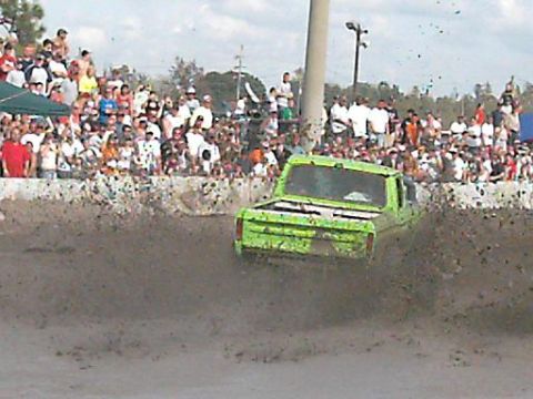 1986 Ford Bronco - A green pickup truck navigates a mud pit during a competition, with a large crowd watching from the sidelines.