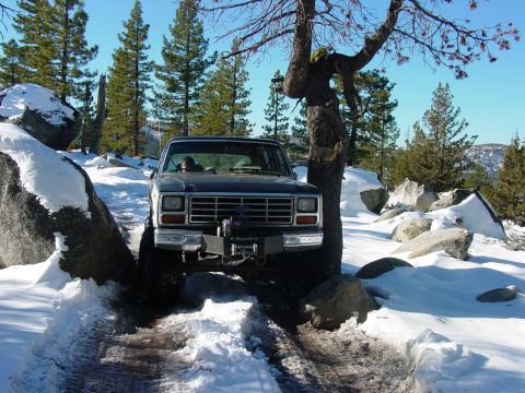 1986 Ford Bronco - Ford Bronco navigates a snowy mountain path with large rocks and trees.