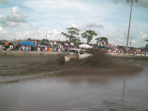1986 Ford Bronco - A white pickup truck navigates a mud pit, sending up a large spray of mud as onlookers watch.
