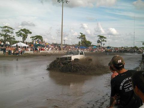 1986 Ford Bronco - A white pickup truck navigates a muddy track during a competition, with a large crowd gathered to watch.