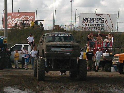 1986 Ford Bronco - Mud bog truck with custom windshield text sits in the mud, surrounded by spectators and other vehicles.
