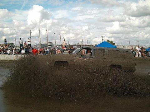 1986 Ford Bronco - A driver navigates a muddy track, sending a massive plume of dirt into the air as onlookers observe.
