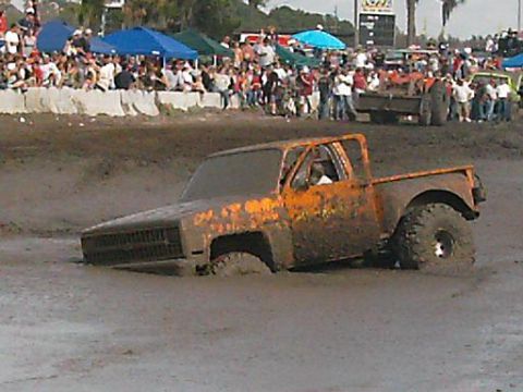 1986 Ford Bronco - A muddy pickup truck is stuck in a mud pit with spectators in the background.