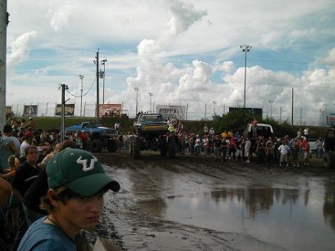 1986 Ford Bronco - Monster trucks compete in a mud pit event with a crowd of onlookers gathered around.