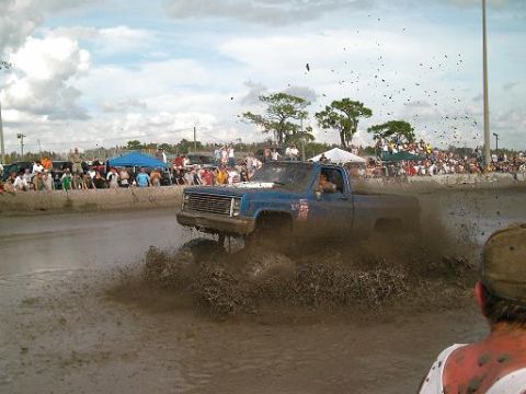 1986 Ford Bronco - A blue pickup truck navigates a mud bog with a crowd watching in the background.