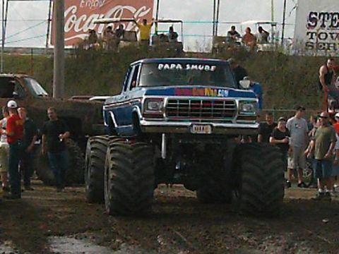 1986 Ford Bronco - A modified truck with oversized tires sits in the mud, with spectators and another vehicle nearby.