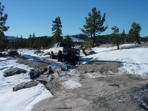 1986 Ford Bronco - A white SUV tackles a challenging off-road path covered in snow and rocks.