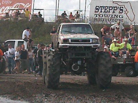 1986 Ford Bronco - Monster truck event with a lifted pickup and spectators gathered around.