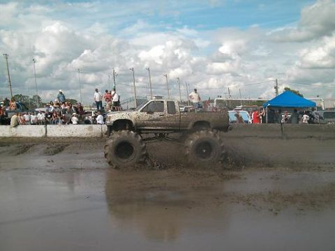 1986 Ford Bronco - A lifted truck with large tires navigates a muddy track during an event, kicking up spray.