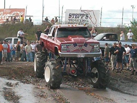 1986 Ford Bronco - A modified red pickup truck with oversized tires navigates a muddy track in front of a crowd.
