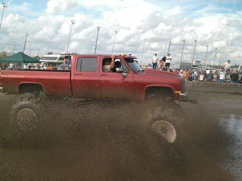 1986 Ford Bronco - Modified pickup truck powers through a mud pit with spectators watching.