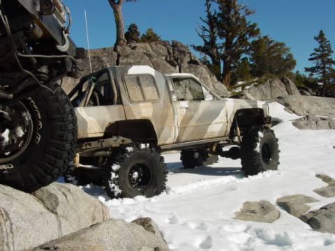 1986 Ford Bronco - A heavily modified truck with oversized tires navigates a snowy, rocky terrain under a clear blue sky.