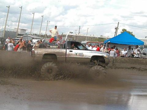 1986 Ford Bronco - A lifted pickup truck navigates a muddy track during a competition, with spectators watching from the sidelines.