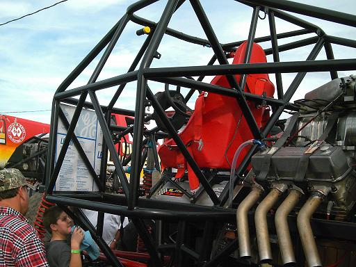 Roll cage inside a custom vehicle with a red seat and exposed engine with exhaust pipes. Another vehicle is visible to the left