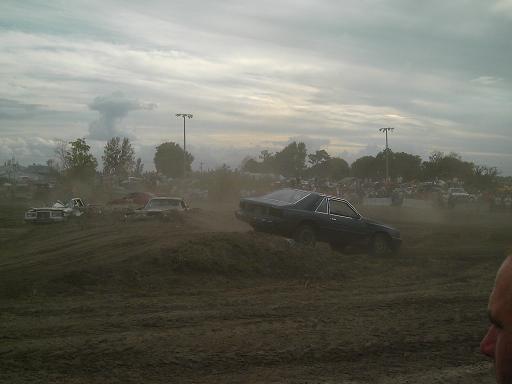 Several cars are in a dusty field with a crowd in the background