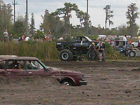 1986 Ford Bronco - A muddy scene with a maroon car partially submerged in mud and a large lifted black truck nearby.