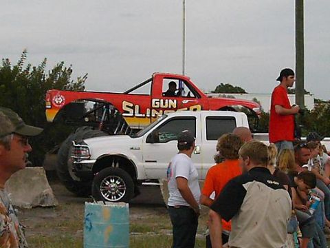 1986 Ford Bronco - A crowd watches a red monster truck, labeled "GUN SLINGER," positioned over a white pickup truck at an event.