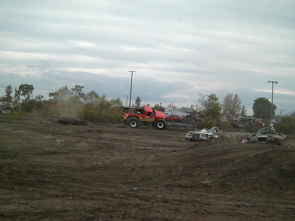 Red monster truck with 'SLIMER' on the side driving in a muddy field with other wrecked cars