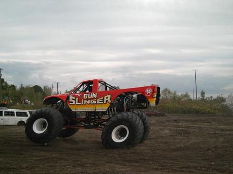 1986 Ford Bronco - A monster truck named 'GUN SLINGER' sits on a dirt lot under a cloudy sky, with a white van visible in the background.