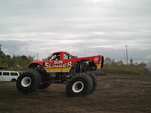 Red monster truck with 'GUN SLINGER' text, large white-rimmed tires, and exposed suspension, parked on dirt