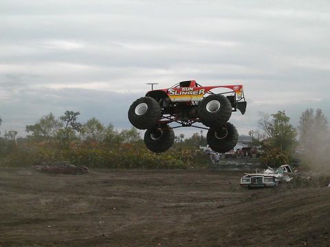 1986 Ford Bronco - The 'Gun Slinger' monster truck soars through the air during an event, with other vehicles visible on the ground below.