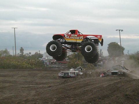 1986 Ford Bronco - A monster truck soars through the air over a pile of wrecked cars during an event.