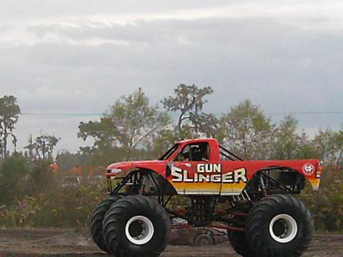 1986 Ford Bronco - A red monster truck named "GUN SLINGER" sits on a dirt track under a cloudy sky.
