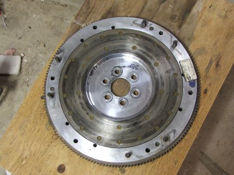 1965 Ford Mustang - A metal flywheel with a toothed outer edge and bolt holes sits on a wooden plank.