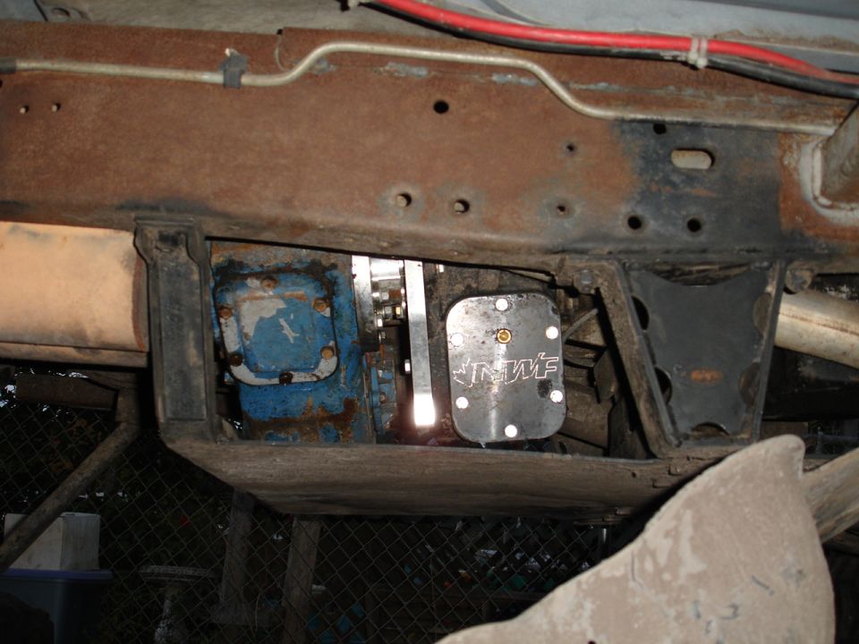 Undercarriage of a rusty vehicle showing drivetrain components and a metal plate with 'INWF' logo