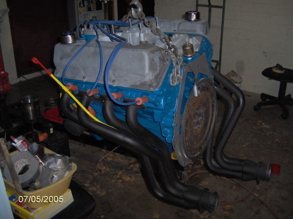V8 engine with blue block, silver valve covers, and black headers, suspended by a chain