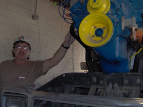 1978 Ford Bronco - Man works on a blue engine with yellow pulleys. The vehicle's front end is visible below.