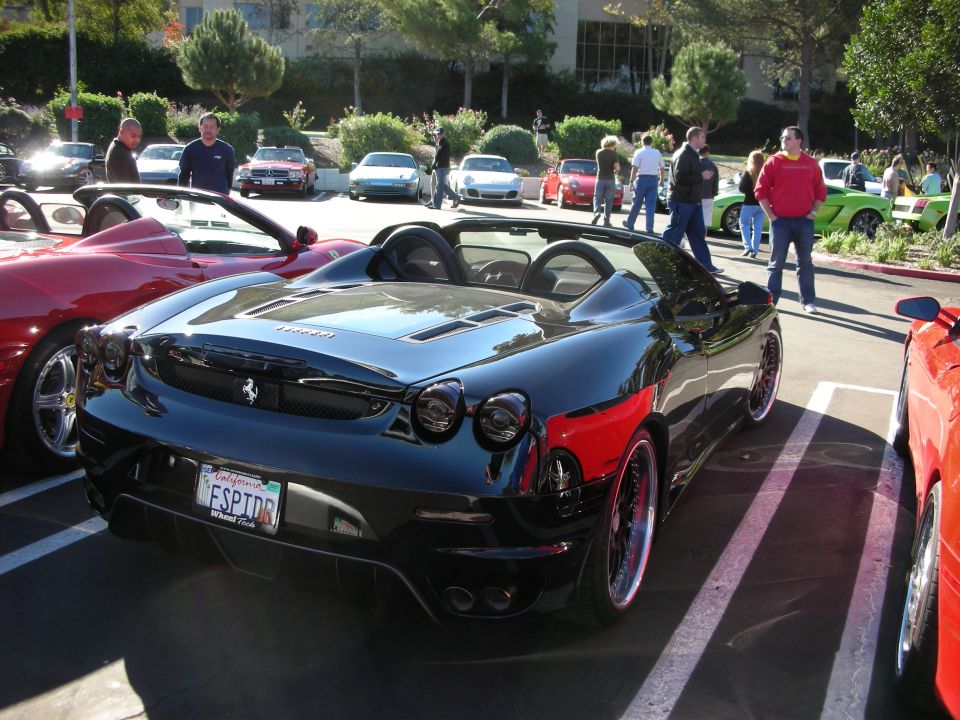 Black Ferrari F430 Spider parked next to a red car, with other vehicles and people in background