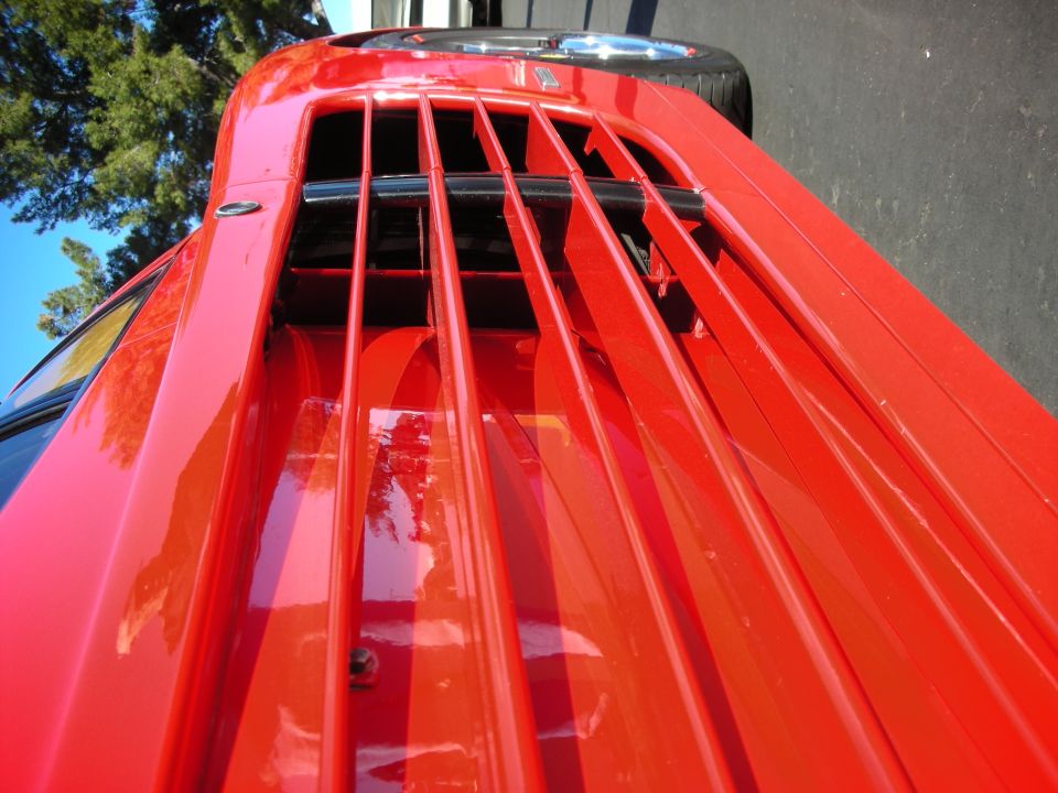 Red sports car rear decklid with prominent horizontal louvers and a central black trim piece. A tire is visible at the top