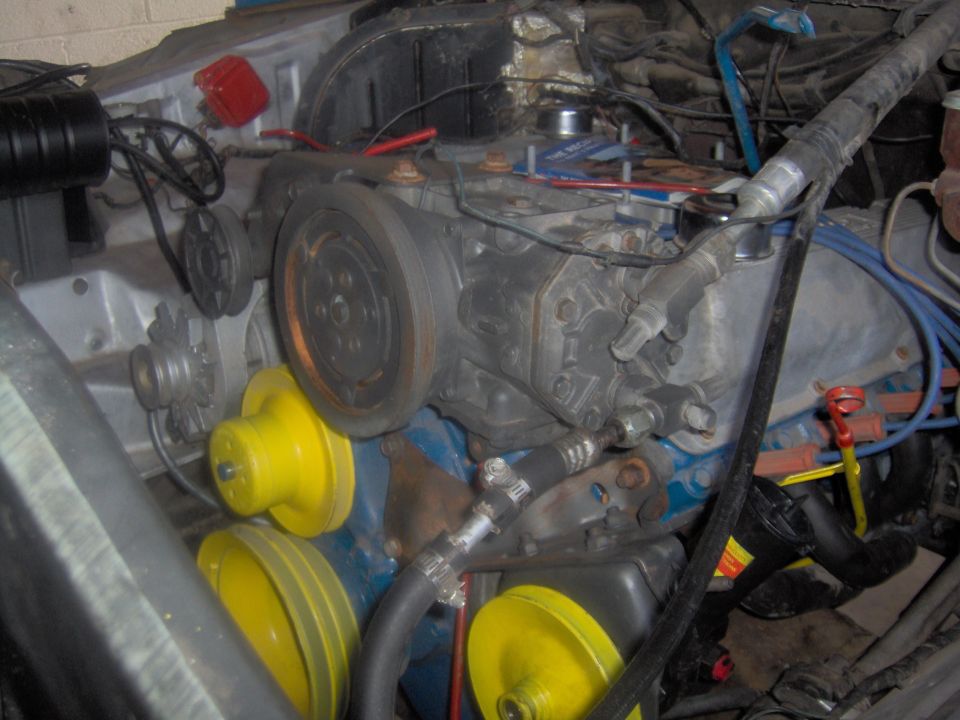 Engine bay showing V-belts, pulleys, and engine components. Yellow covers are visible on some pulleys