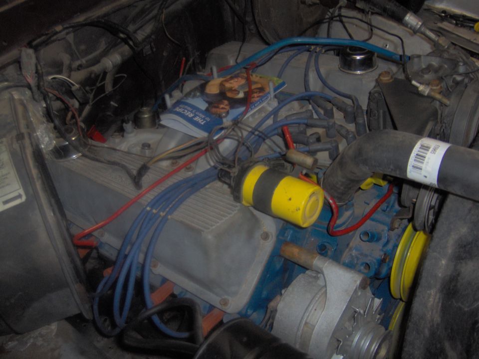 Engine bay with blue spark plug wires, a yellow ignition coil, and a magazine on top of the engine