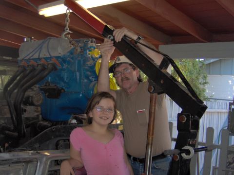 1978 Ford Bronco - Engine hoist lifting a blue engine, with a man and a girl posing for the photo.