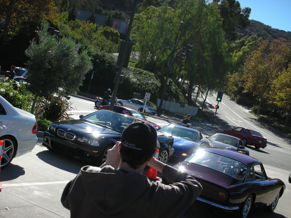 Several cars, including a black BMW, a blue Chevrolet Corvette, and a purple coupe, stopped on a street with a person in the foreground taking a photo