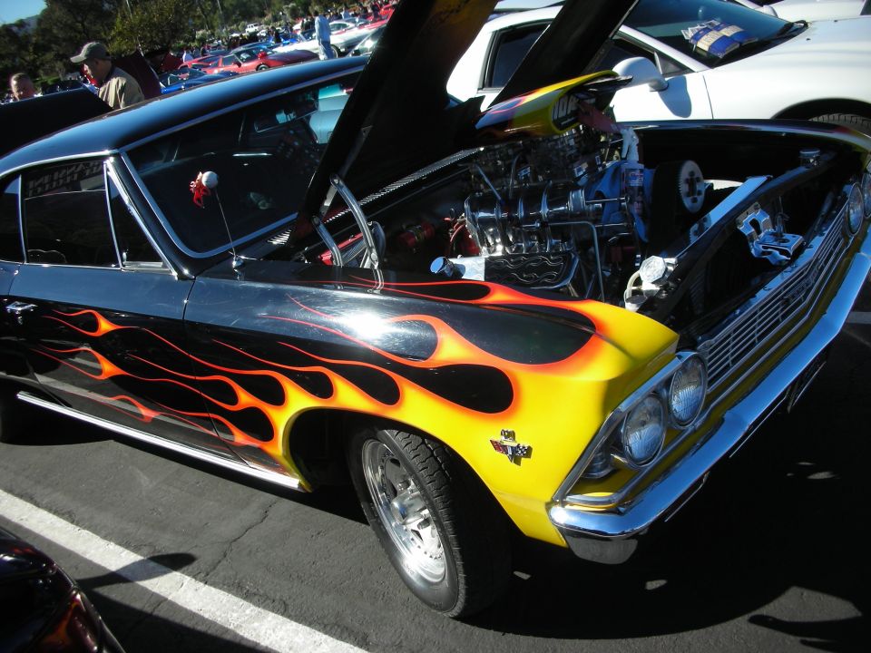 Front quarter view of a black and yellow classic car with flame graphics and an open hood revealing a V8 engine. Other cars are visible in the background