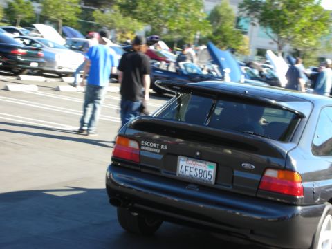 1992 Ford Crown Victoria - A dark colored Ford Escort RS is parked in a lot with other cars and people milling around.