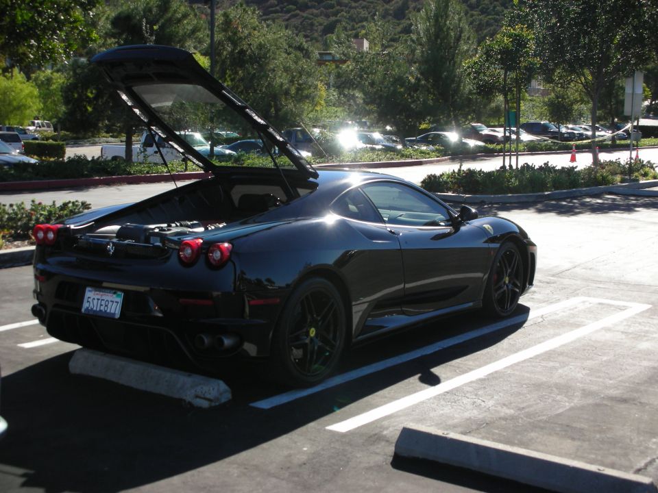 Black Ferrari parked in a lot, rear view with trunk open revealing engine components