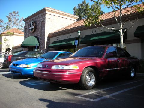1992 Ford Crown Victoria - Two sedans, a red Ford Crown Victoria and a blue Ford Mustang, are parked outside a building.