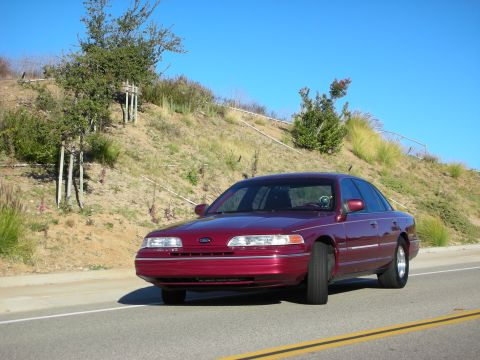 1992 Ford Crown Victoria - A maroon 1992 Ford Crown Victoria sedan is captured in a front 3/4 view on a sunny day, with a natural hillside behind it.