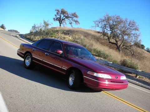 1992 Ford Crown Victoria - A maroon 1992 Ford Crown Victoria cruises along a scenic, winding road under a clear blue sky.
