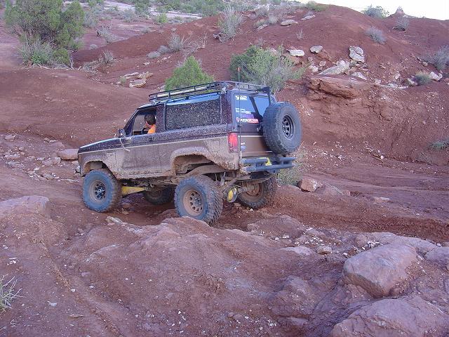 Muddy 1986 Ford Bronco II climbing a rocky trail with a spare tire on the back