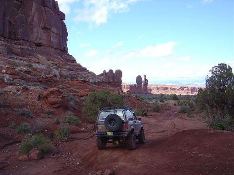 1986 Ford Bronco II - A Ford Bronco II navigates a dirt trail through a desert landscape with distinctive rock formations under a blue sky.