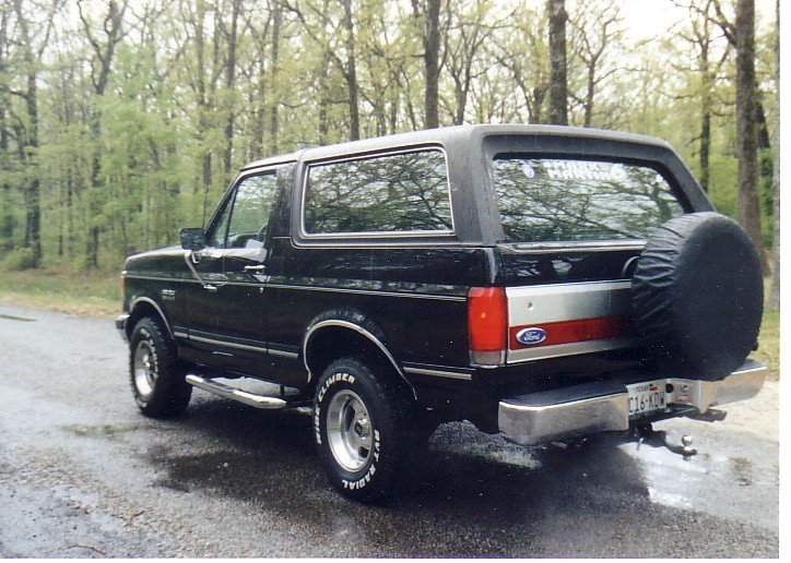 Black Ford Bronco SUV, rear 3/4 view, with spare tire cover, parked on wet road in woods