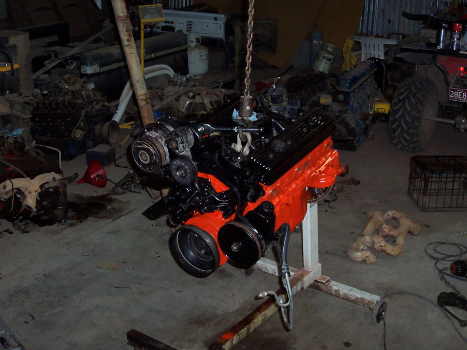 Orange V8 engine with black accessories on a stand in a cluttered garage