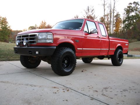 1995 Ford F-150 - Three-quarter front view of a lifted red Ford F-150 XLT extended-cab 4x4 on a concrete driveway, fitted with a black tube-style front bumpe…