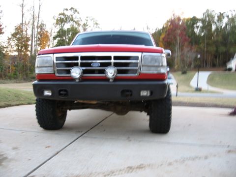 1995 Ford F-150 - Front-on shot of a lifted red Ford pickup parked on a concrete driveway, with chrome bar grille and Ford oval, two round auxiliary driving…