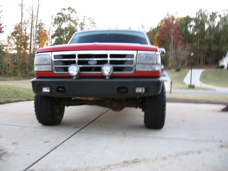 Lifted red Ford pickup head-on with driving lights, black bumper and mud-terrain tires